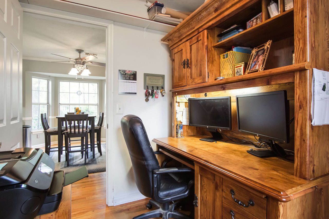 The room features a wooden desk with a computer and bookshelves. The background shows a dining area with a table and chairs.