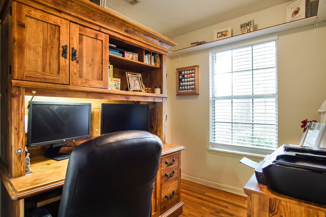 A home office setup with a wooden desk, computer, and bookshelves. The room has a window letting in natural light, and the desk is organized and functional.