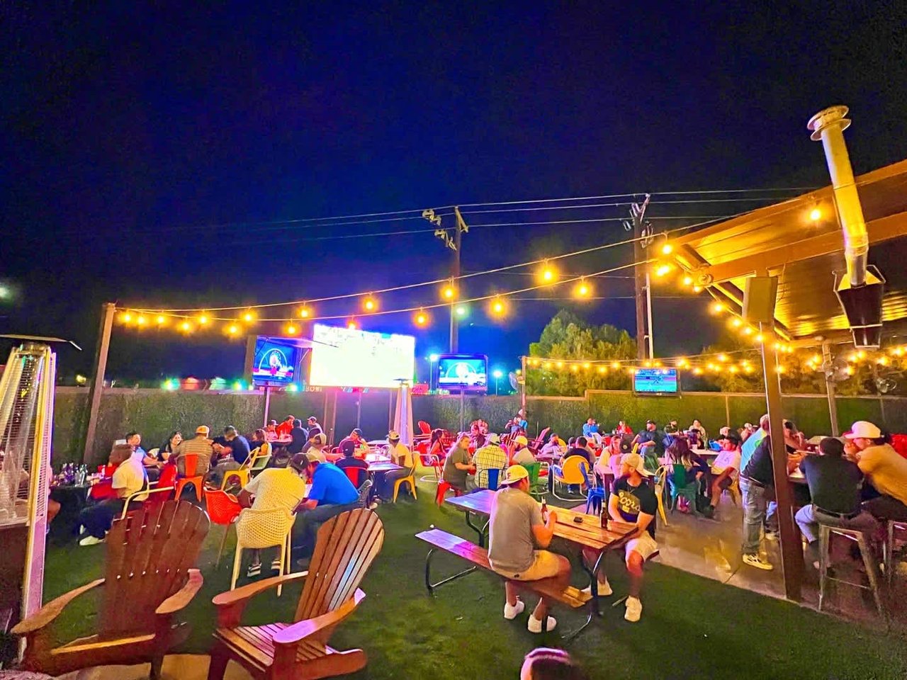Crowded outdoor patio at night with string lights, picnic tables, and large screens.