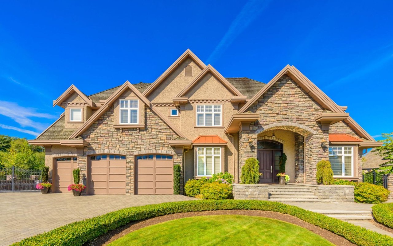 A grand Colonial-style brickhouse with a lush front lawn and hedges, patio, spacious driveway, and expansive two-car garage.