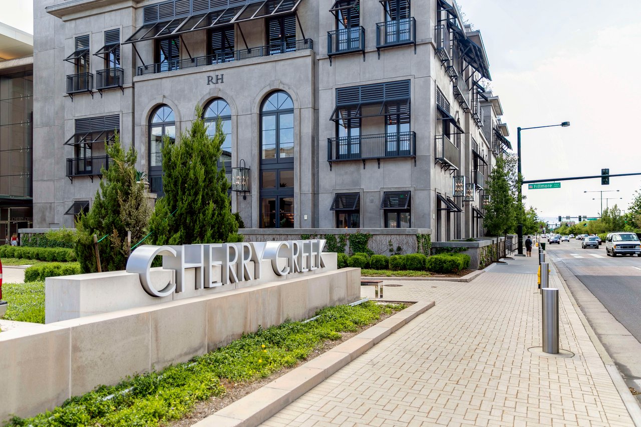 A sign that reads "Cherry Creek" in front of a building, the sign is located on a sidewalk with a metal railing.