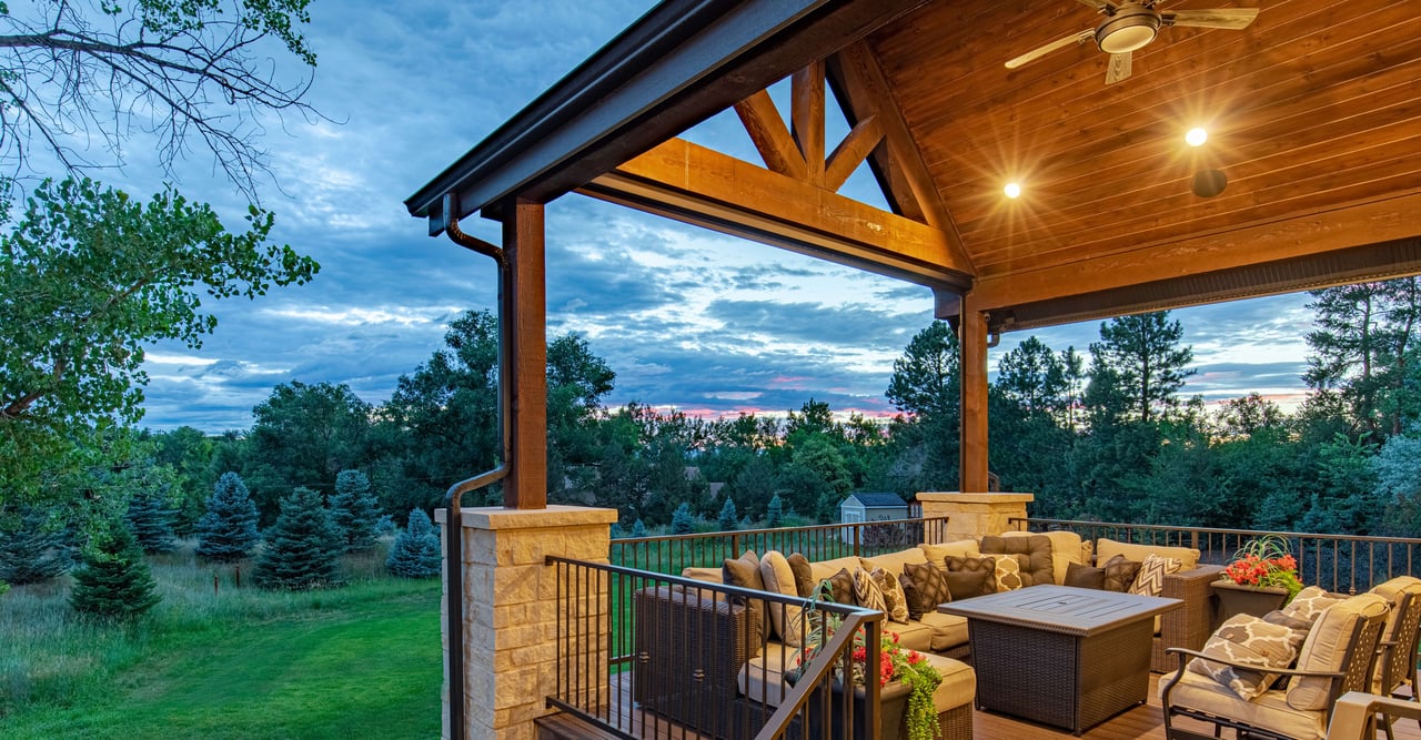 A cozy covered deck with a couch, chairs, and a table. The deck is surrounded by trees and greenery