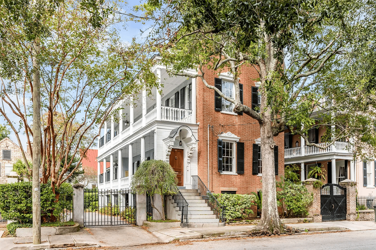 Red brick home with white columned porches, black shutters, and mature trees along the street.