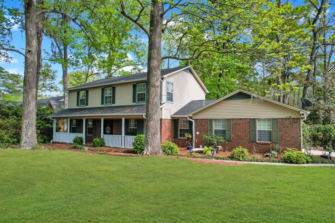 Another exterior view of a two-story house with a large front porch. The house is surrounded by well-maintained landscaping and tall trees.