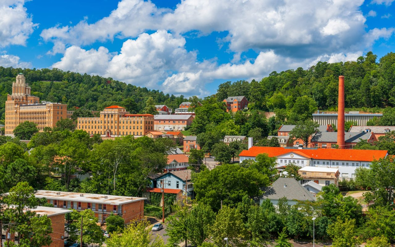  A vibrant aerial view of a town nestled in a valley surrounded by lush green, tree-covered hills under a bright blue sky with fluffy white clouds.