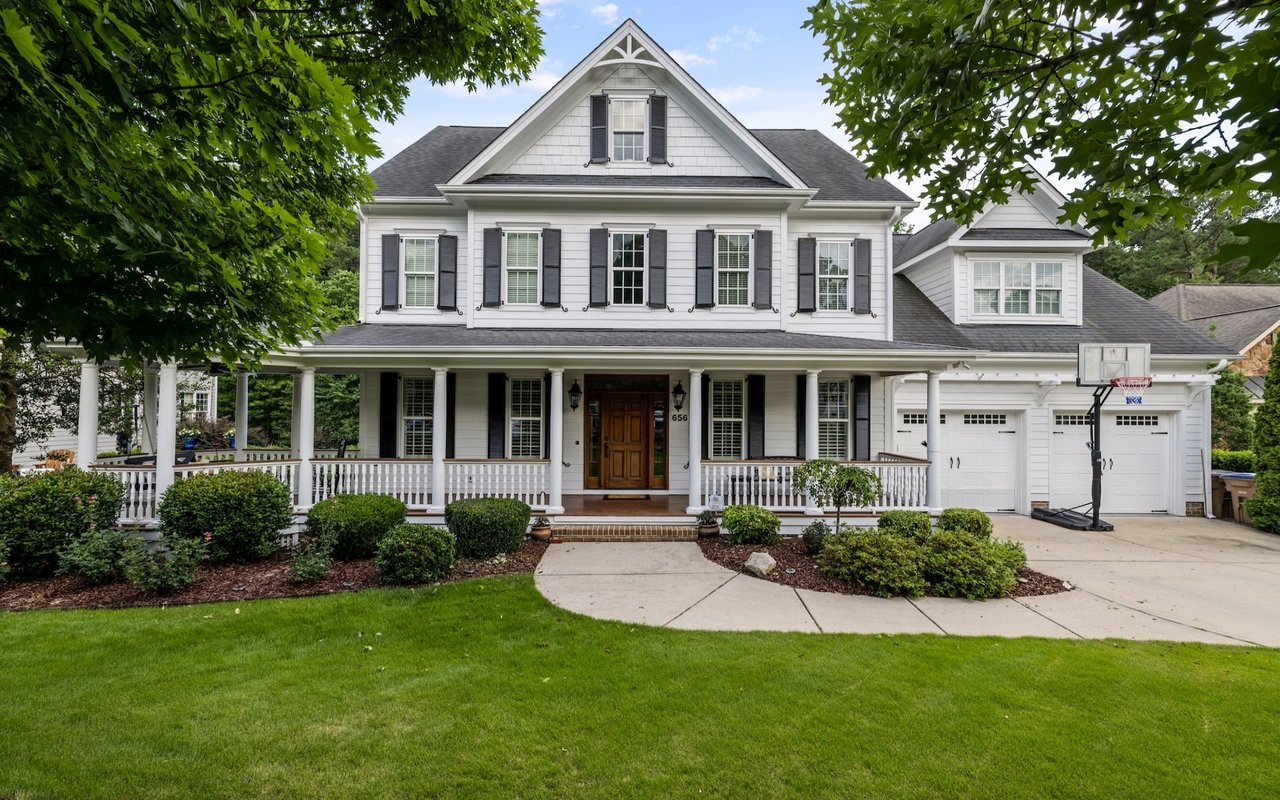 The house is surrounded by trees, and there is a green lawn in the front. The sky is a clear blue, and the sun is shining.