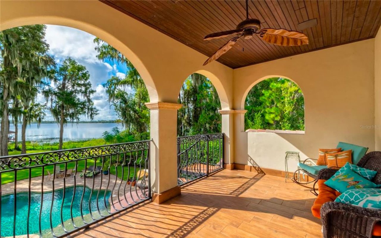 A balcony overlooking a pool and lake, with lounge chairs and a metal railing, surrounded by trees.