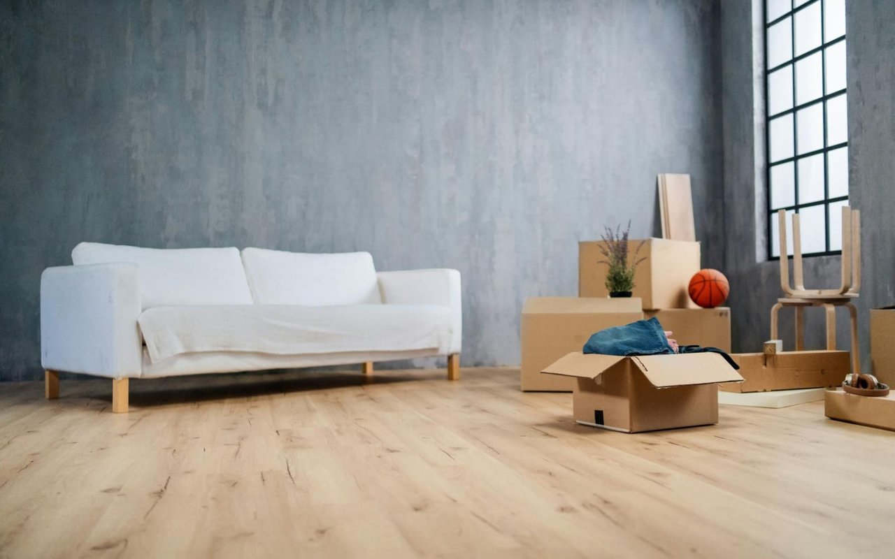 A fairly bare living room with wooden flooring, a two-seater couch, and several cardboard boxes stacked against the walls.