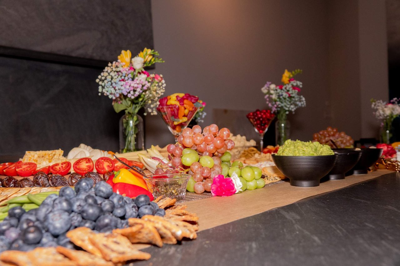Buffet of grapes, blueberries, sliced tomatoes, cheese, and crackers with flower arrangements behind it.