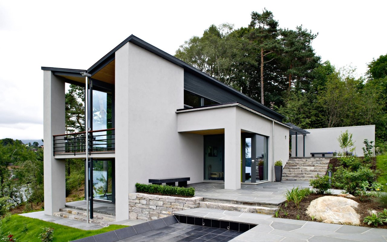 Modern white home with dark rooflines, large windows, stone patio, and landscaped grounds.