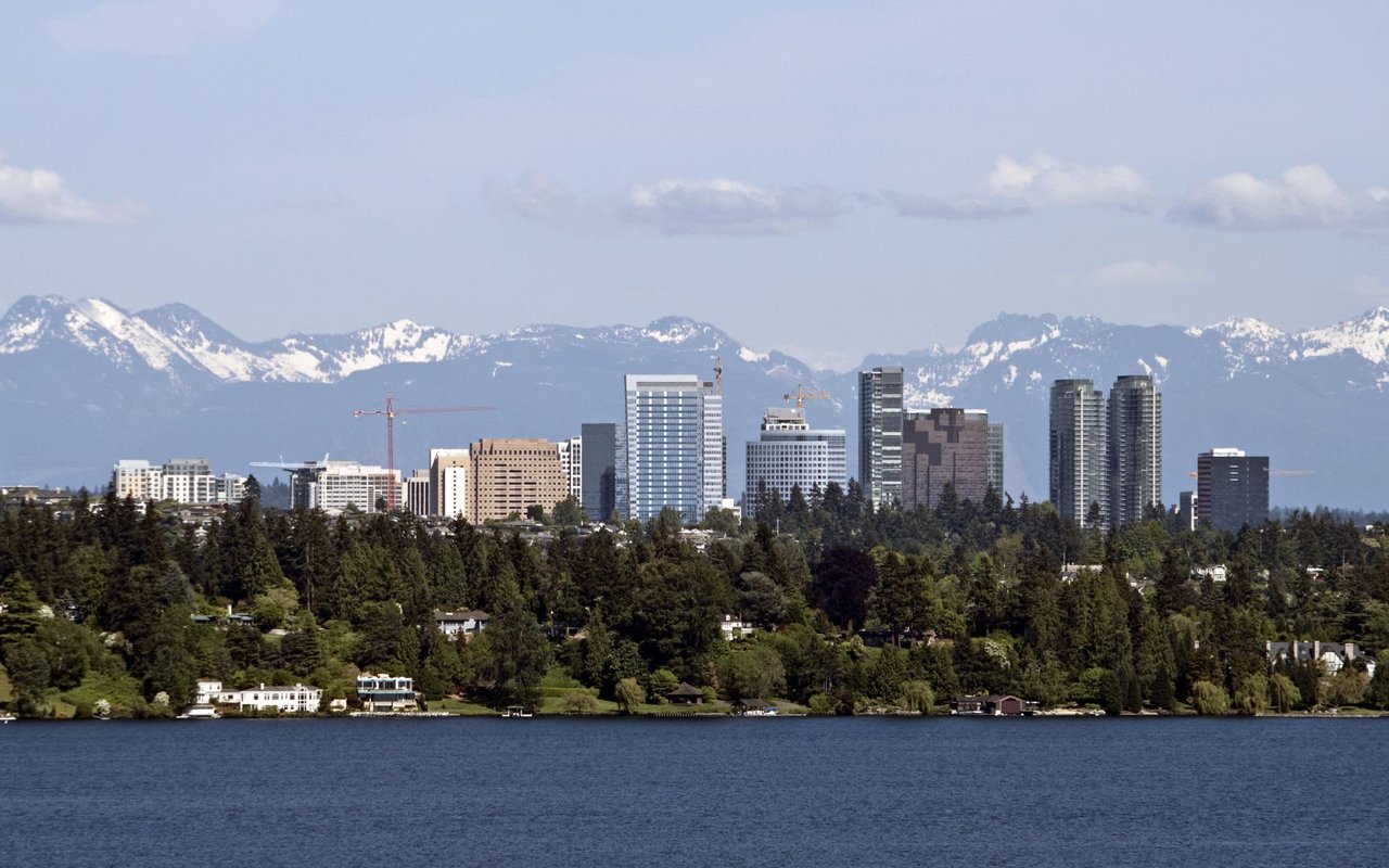 A panoramic view of a city skyline across a wide body of water, with snow-capped mountains in the distant background under a partly cloudy sky.