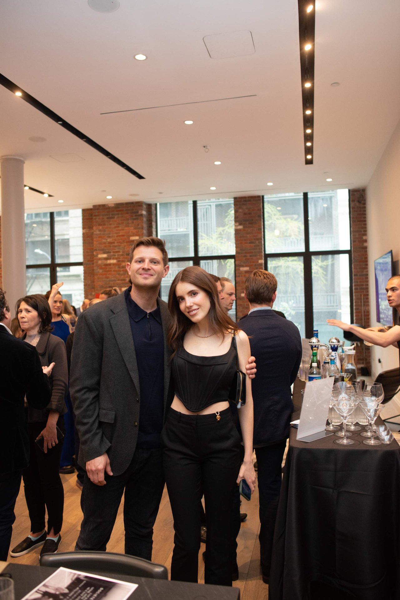 Guests pose together at an indoor event, with a drinks table and other attendees nearby.