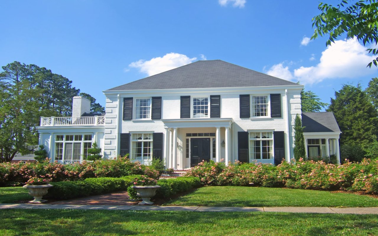 A large two-storey white house with black shutters with a lush green yard in front of the house and a clear blue sky above it.