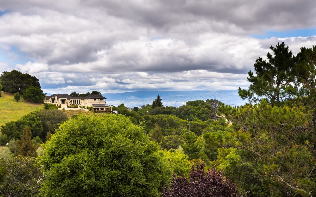 A two-story house with a brown exterior and a black roof on a hill, surrounded by trees and rolling hills in the background.