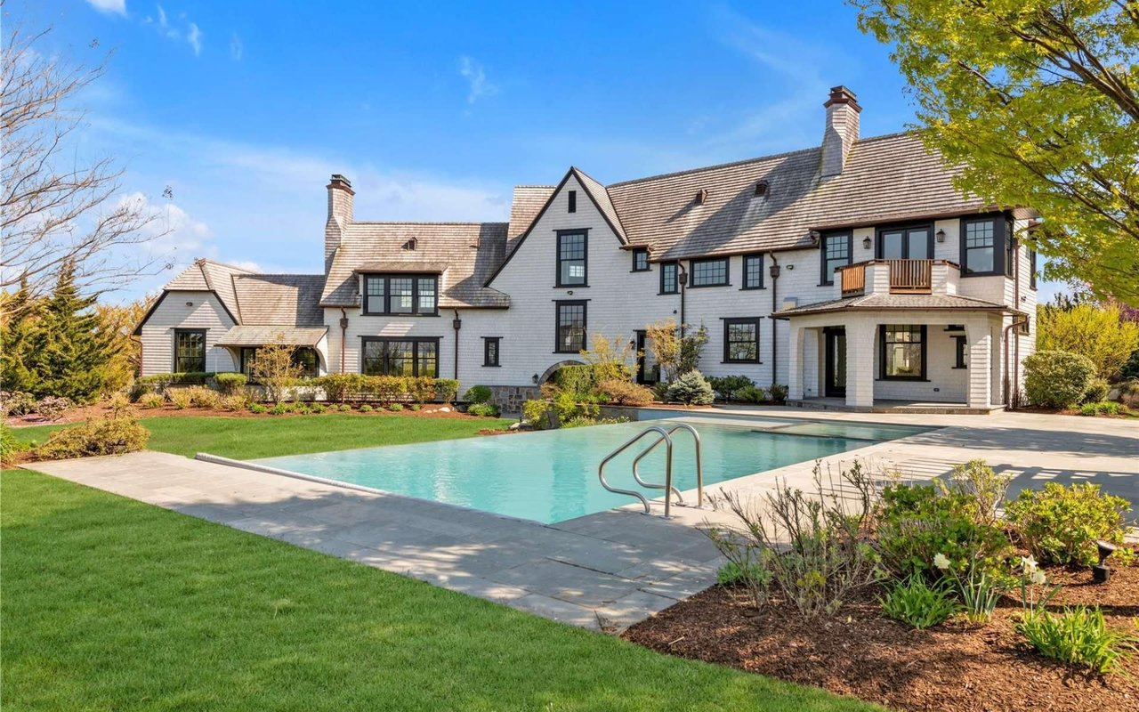 A large white house with black trim and a shingle roof, featuring a pool and expansive lawn.