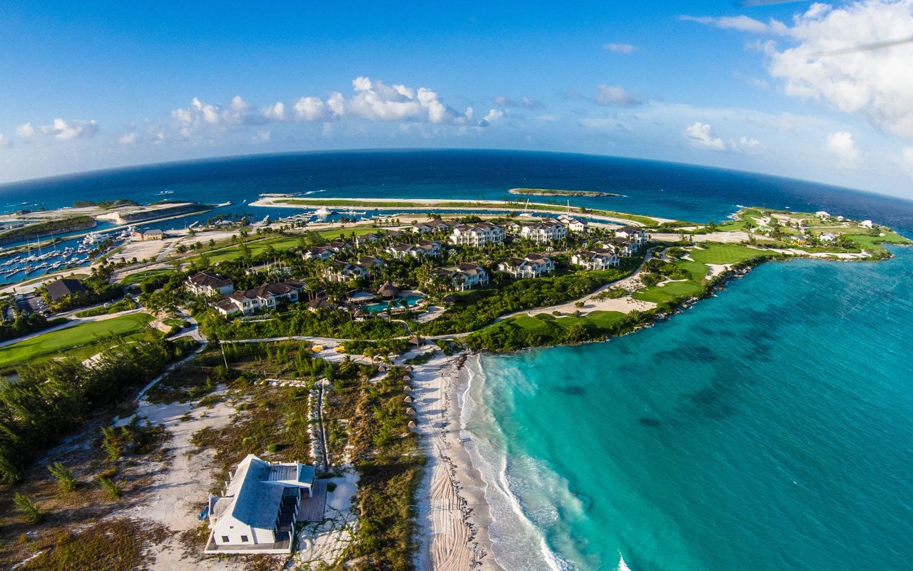 An expansive aerial view, taken with a wide-angle lens, captures a luxurious island resort with white buildings and lush green golf courses