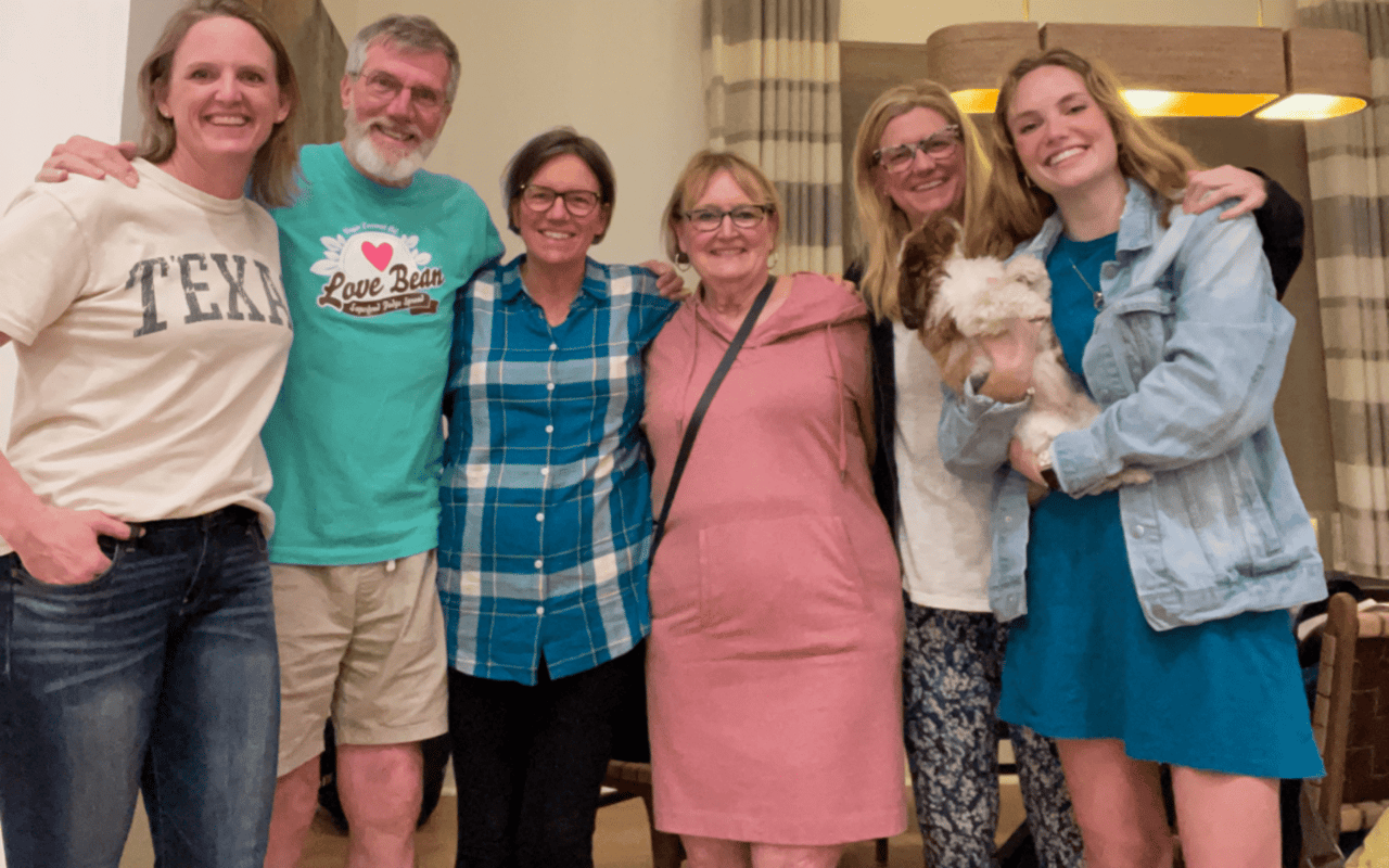  A group of six smiling individuals are standing close together, seemingly posing for a photo in an indoor setting. 