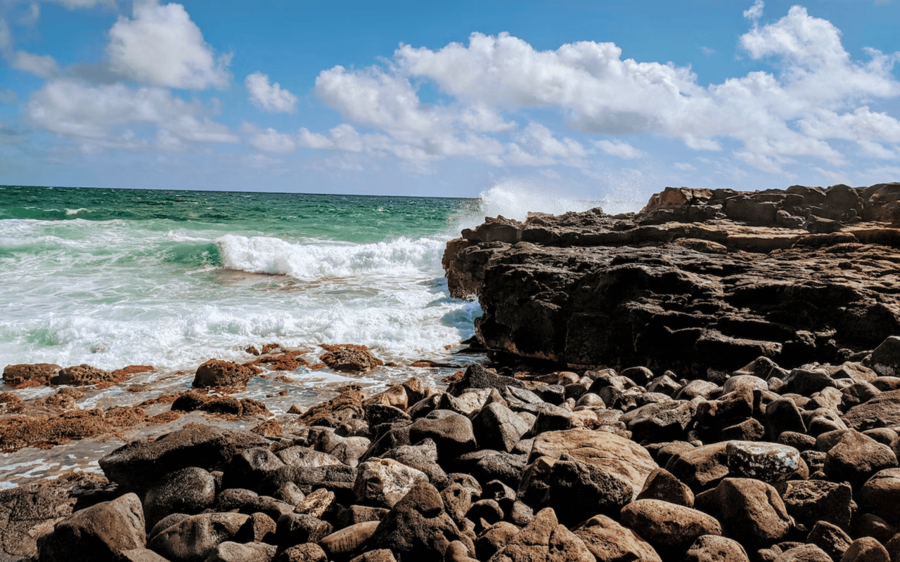 Rocky shoreline, golden sand, white-capped waves, and clear turquoise water of Poipu Beach.