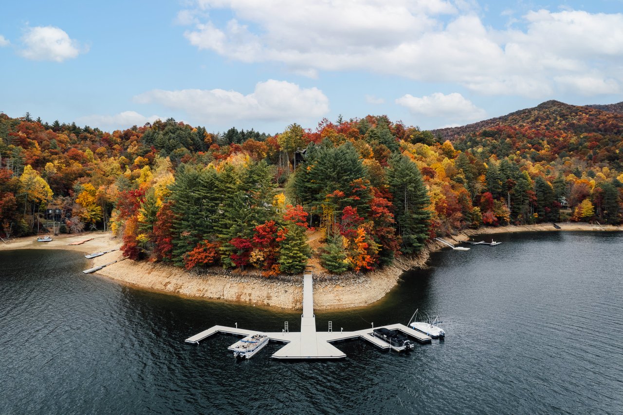 Alt=Aerial view of a small peninsula with a dock platform extending into a lake, surrounded by autumn-colored trees.