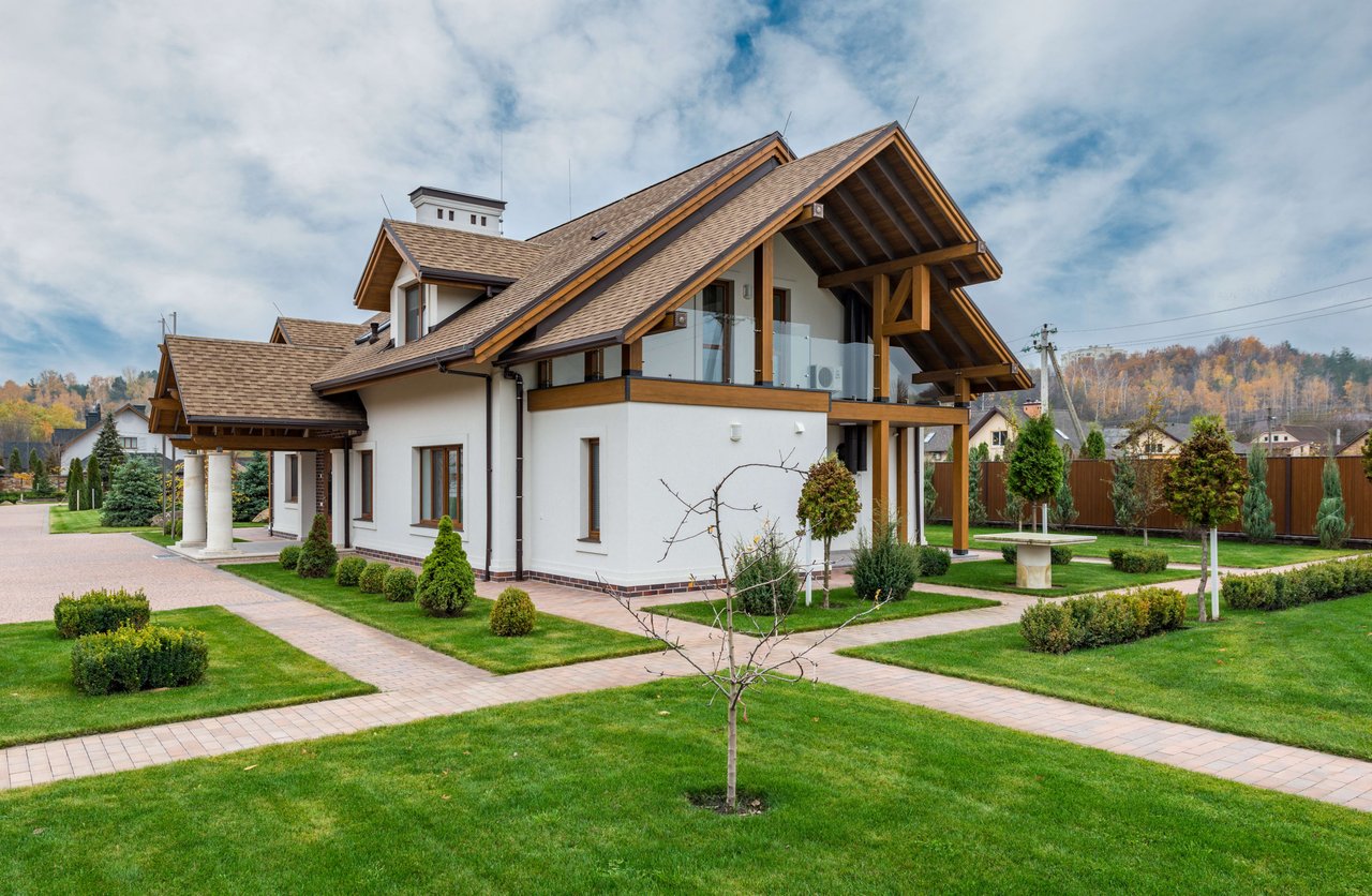 A modern white house with a brown roof and wooden accents sits on a manicured lawn with paved walkways.