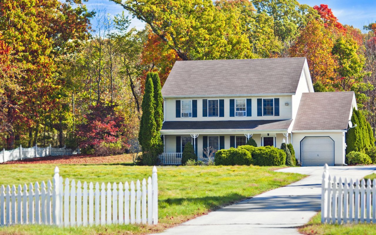 A two-story colonial house with a picket fence, a covered porch, a front yard, and a driveway leading to a two-car garage.