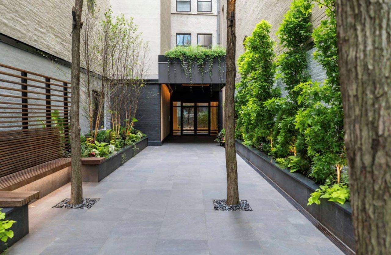 Residential courtyard with leafy planters, mature trees, a wooden bench, and a glass entry.