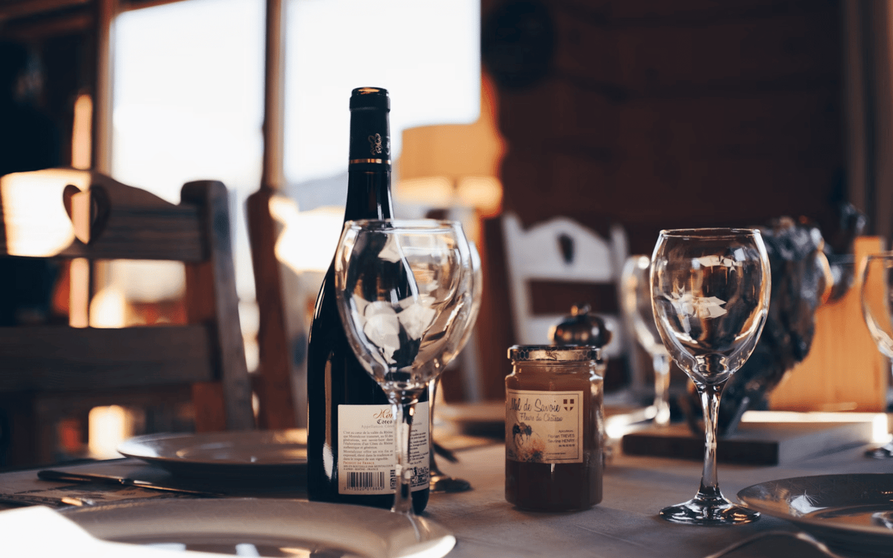 A bottle of wine, two empty glasses, and a jar of honey await on a sunlit table.