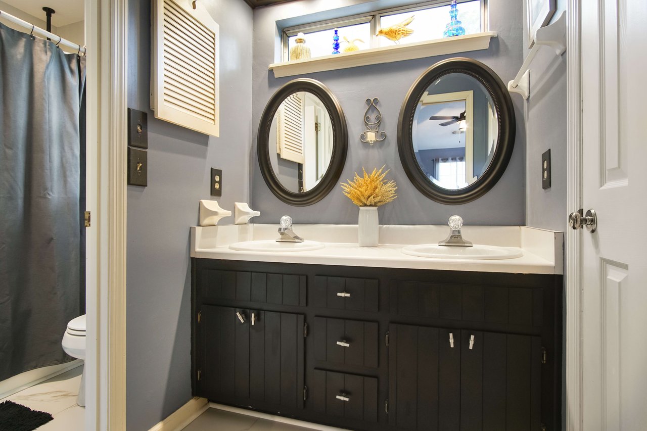 A bathroom with double sinks, each with its own round mirror. The bathroom has dark cabinets, light-colored countertops, and is decorated with modern fixtures.