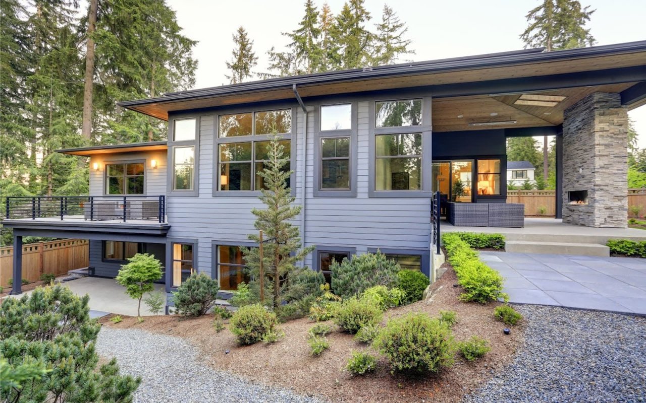  A large, contemporary house with light grey siding and dark gray trim stands amongst tall evergreen trees.