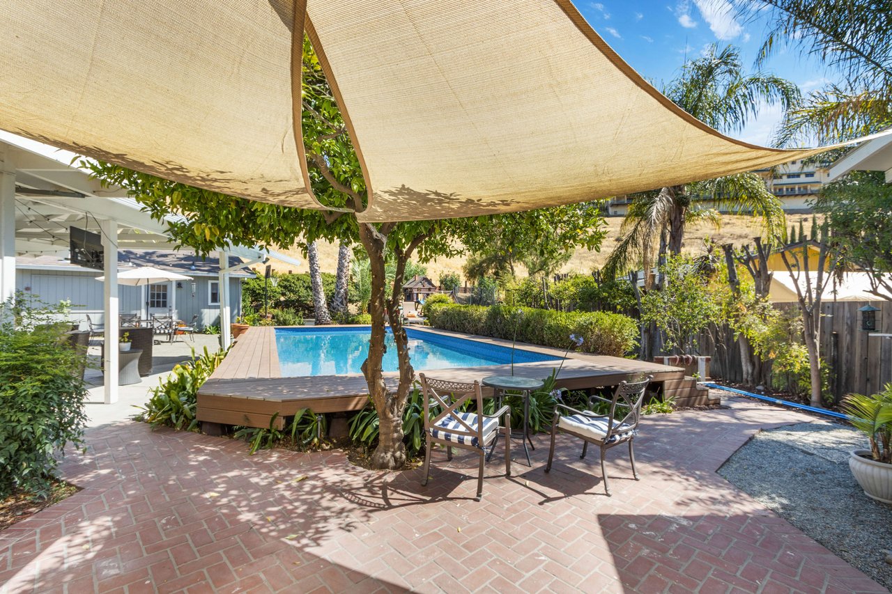 Backyard pool and raised deck beneath large shade sails, with a brick patio and outdoor seating.