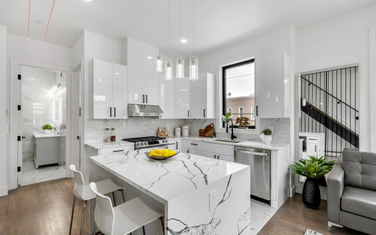 A white kitchen with a marble countertop, stainless steel appliances, white cabinets, and a window above the sink.