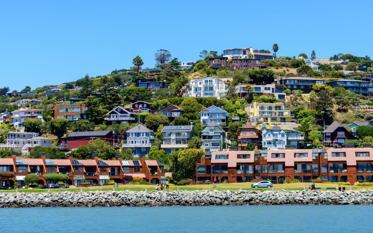 A hillside town with colorful houses overlooks a bay with boats and a distant city skyline.