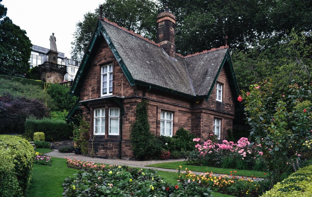  A charming, dark stone cottage with a steep grey roof and dark green trim stands nestled amidst a lush, well-manicured garden. 