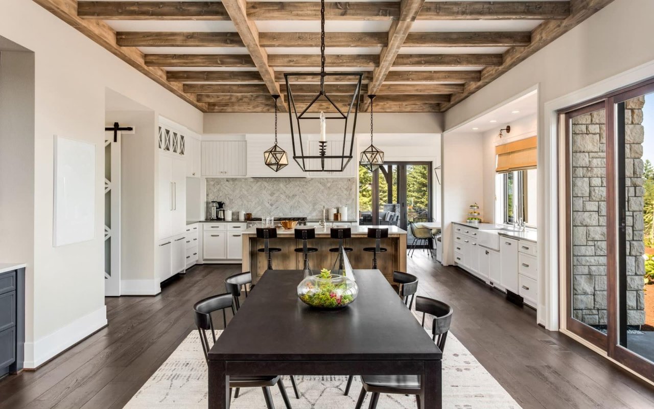 A dining area with light cabinets, a wooden table and chairs, a large window, a wood beam ceiling, and a hardwood floor.