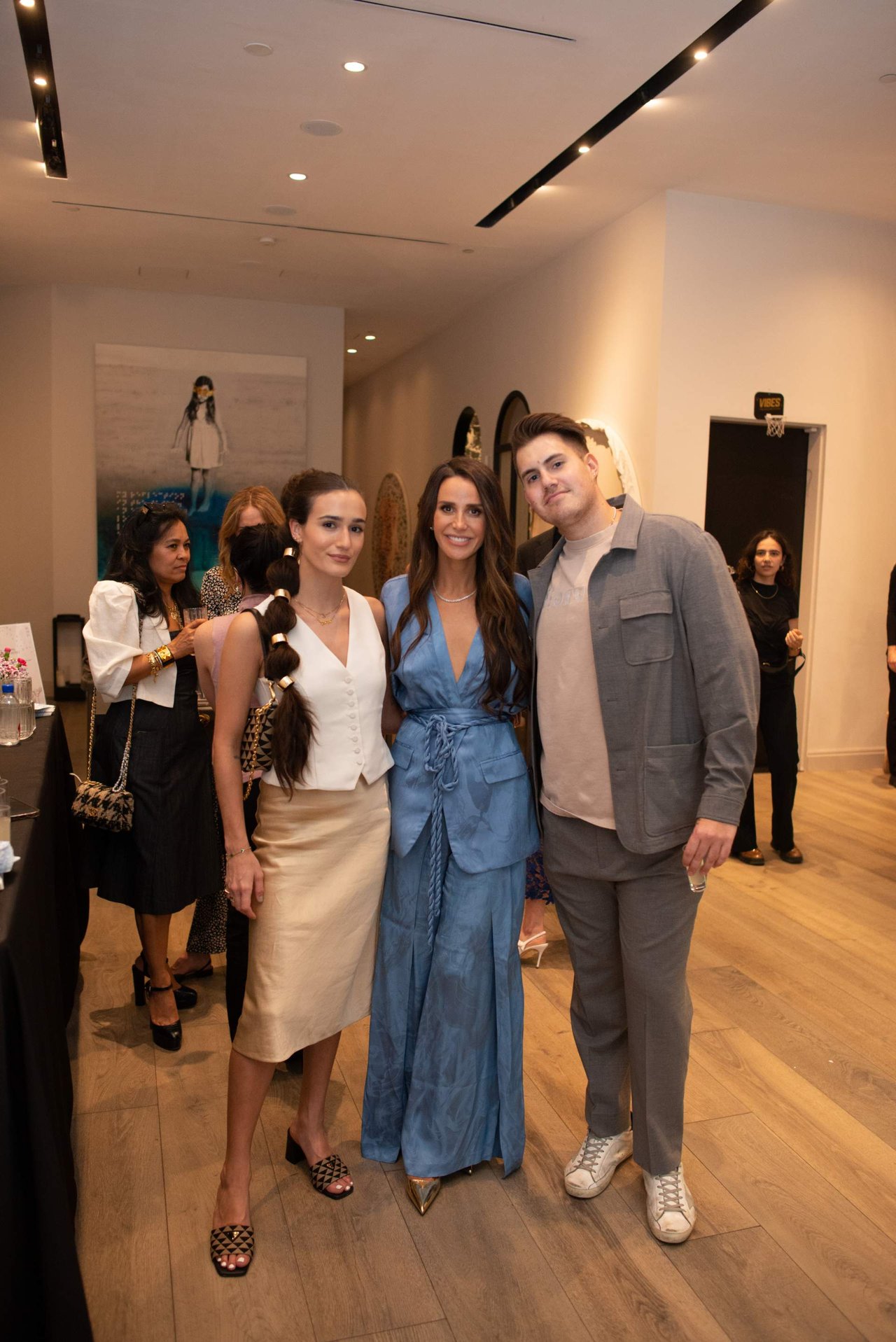 Three event attendees pose in the foreground, dressed in white, blue, and gray, with other guests behind them.