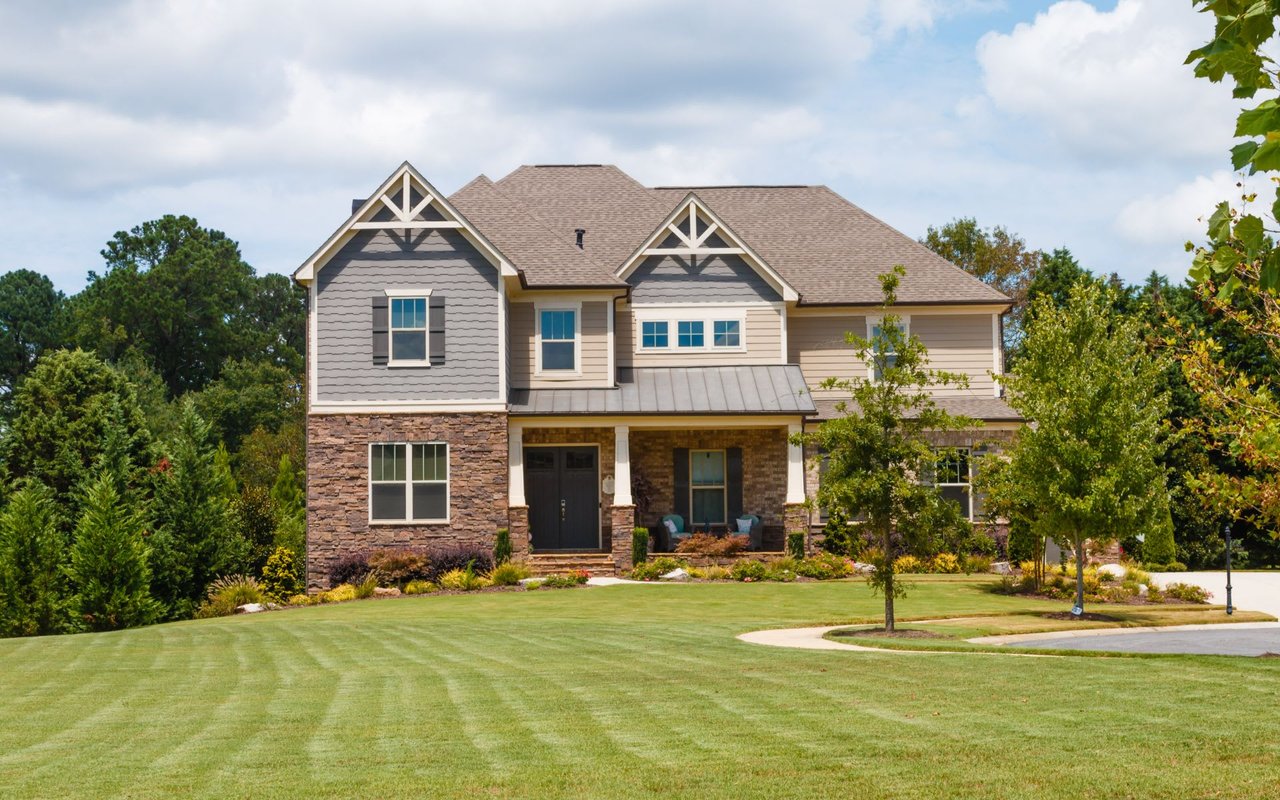 A two-story house with black shutters, a lush green yard, a covered porch, and a stone pathway leading to a backyard patio.