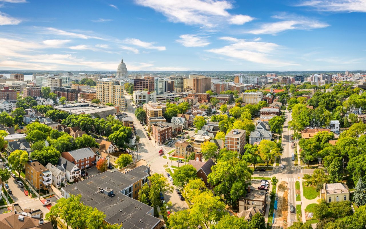 An aerial view of a city skyline with tall buildings, houses of varying styles, and patches of greenery scattered throughout.