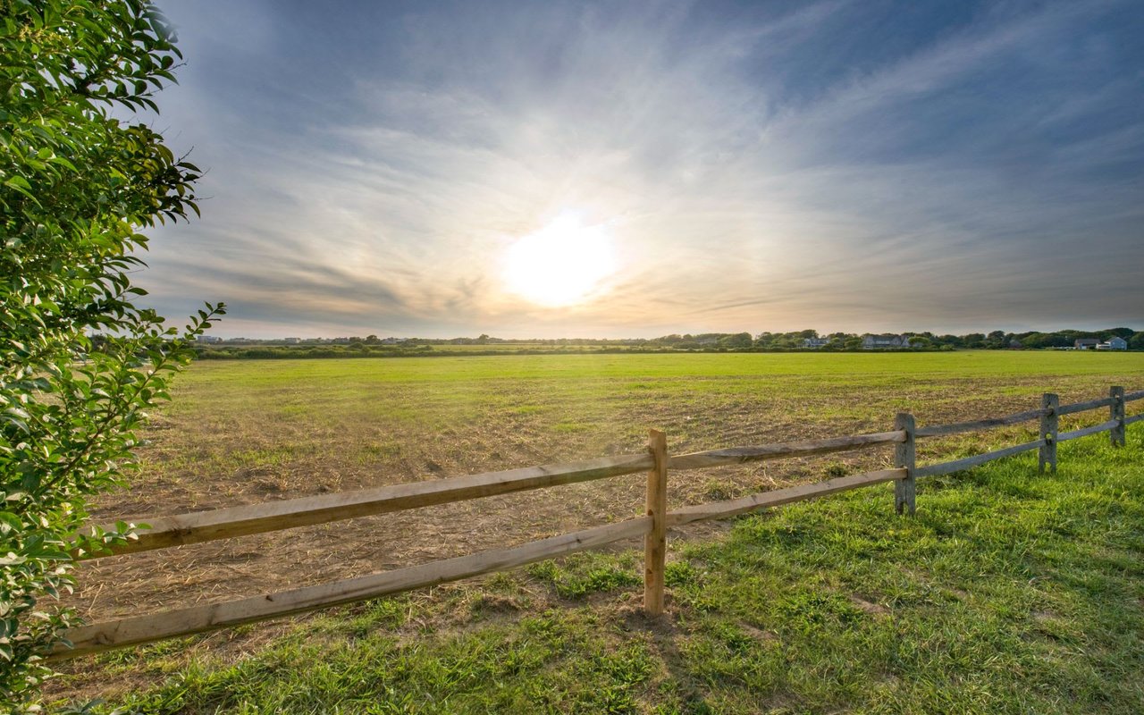 A wooden fence encloses a grassy field under a sunset with orange, pink, and purple hues.