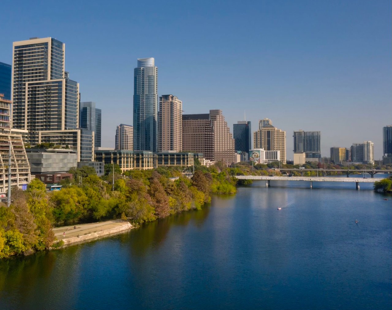 Vibrant cityscape with skyscrapers reflected on a calm river, lush greenery in the foreground.