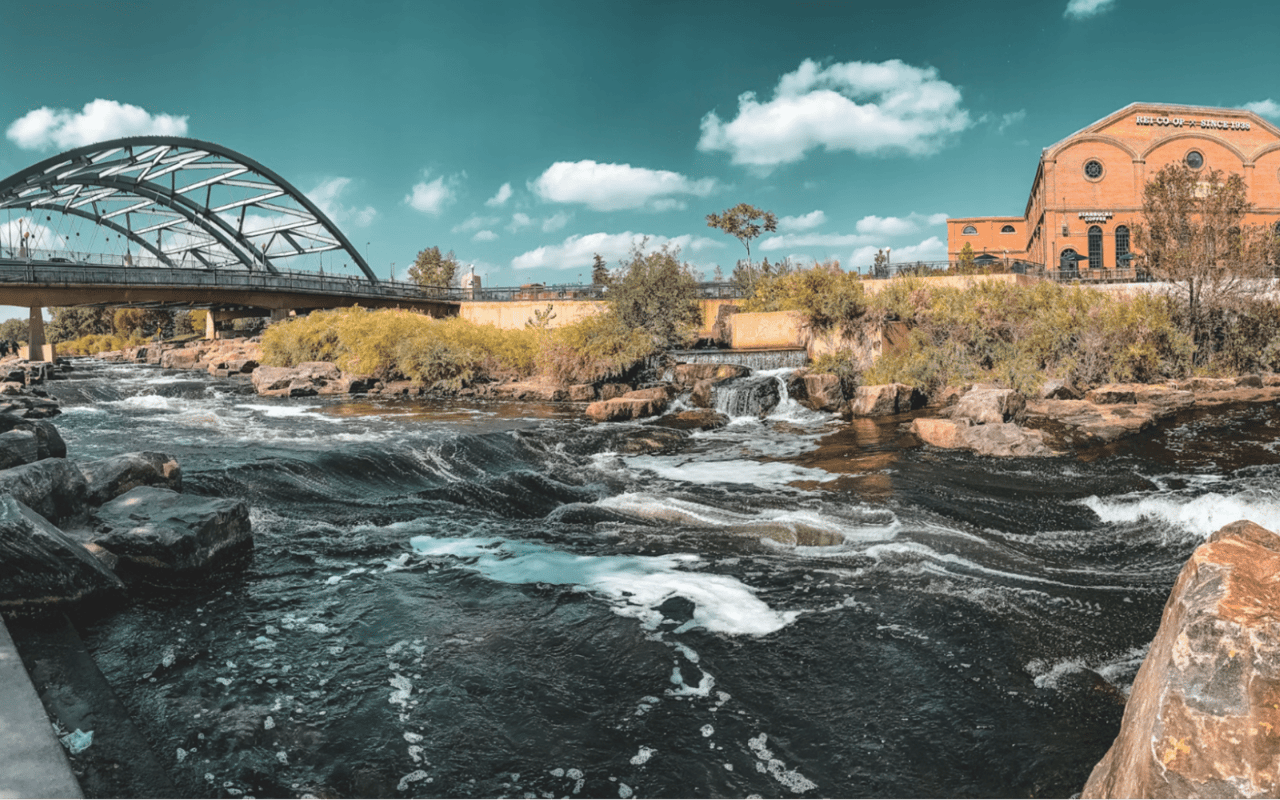  A scenic view captures a wide river with rapidly flowing, dark water, creating white rapids and foam around scattered rocks.