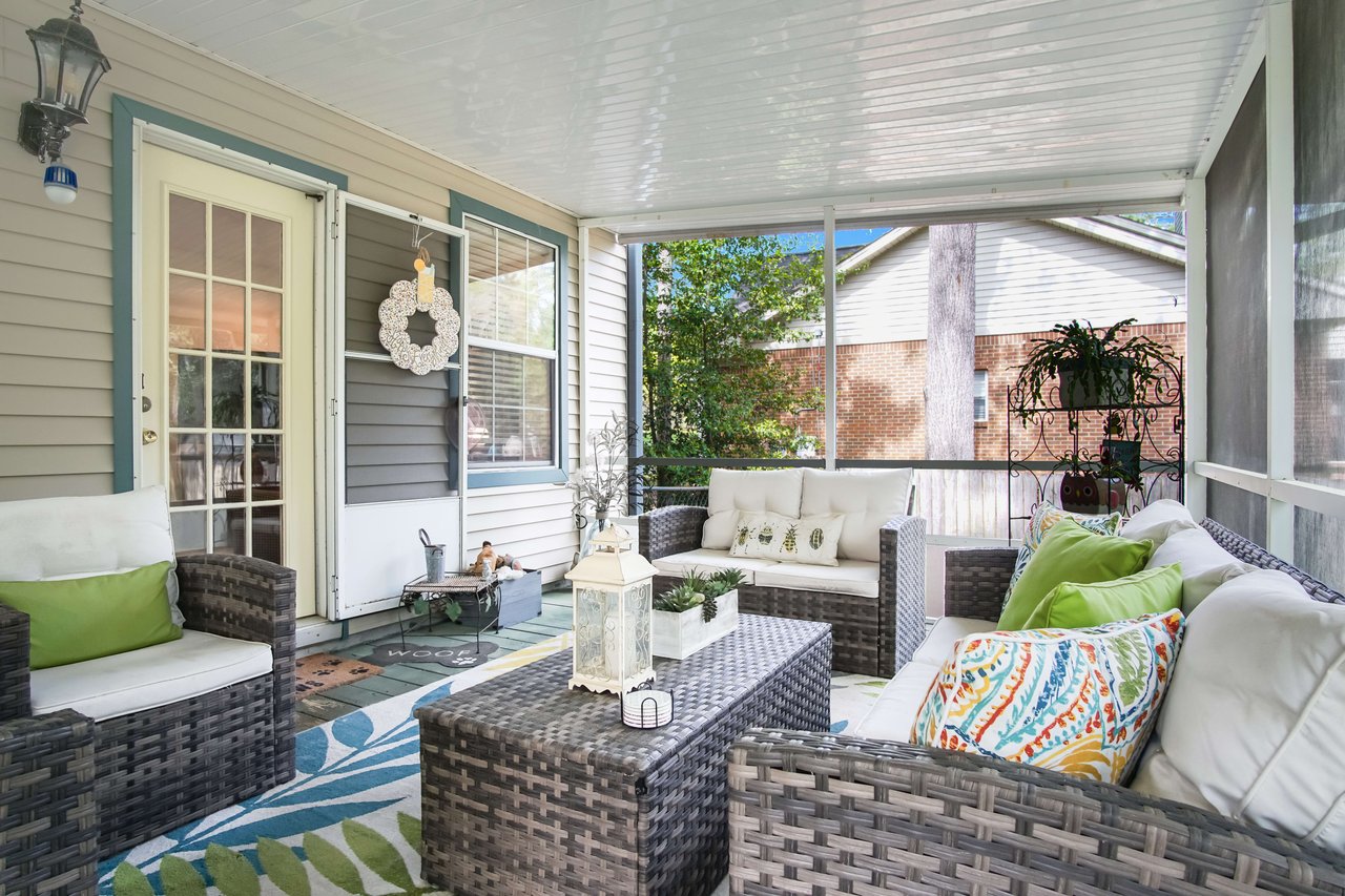 An interior shot of a screened porch or sunroom. The area is furnished with wicker furniture, colorful cushions, and decor, creating a cozy and inviting space for relaxation.