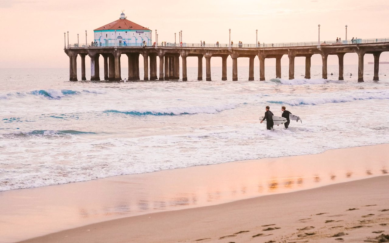 Surfers carry their boards towards the ocean at sunset, with a pier and pavilion in the background.