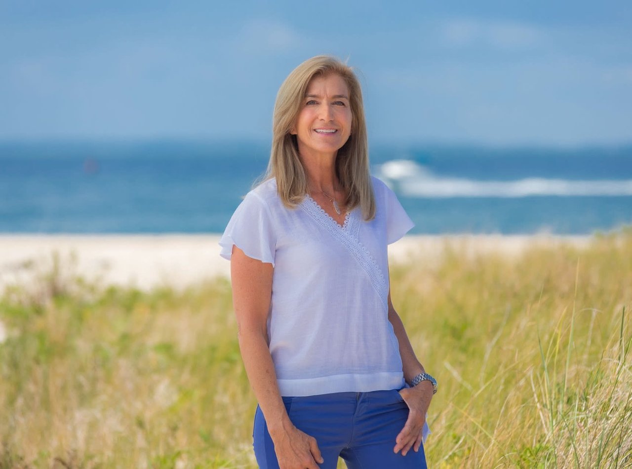 Gina Bradley, licensed real estate professional from The Bradley Team, standing on a sandy beach near ocean waves.