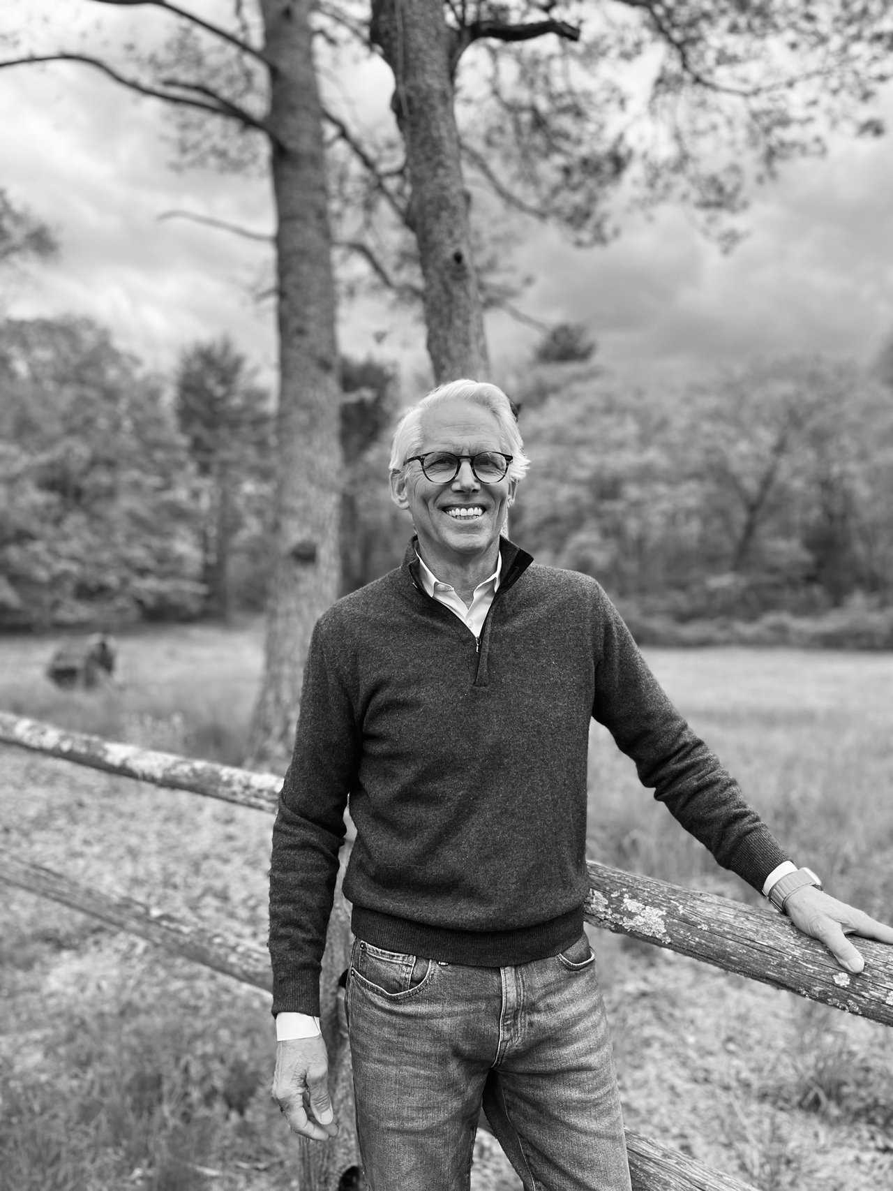 Daniel Ginnel stands outdoors by a wooden fence, smiling. He is wearing glasses, a dark sweater, and jeans. Trees and cloudy skies are in the background. The image is in black and white.