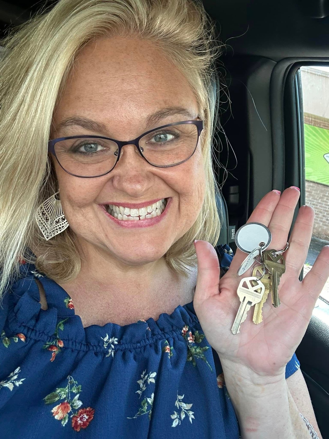 A smiling woman with glasses and a floral top holds up a set of keys inside a car, embodying the confidence of real estate agents.