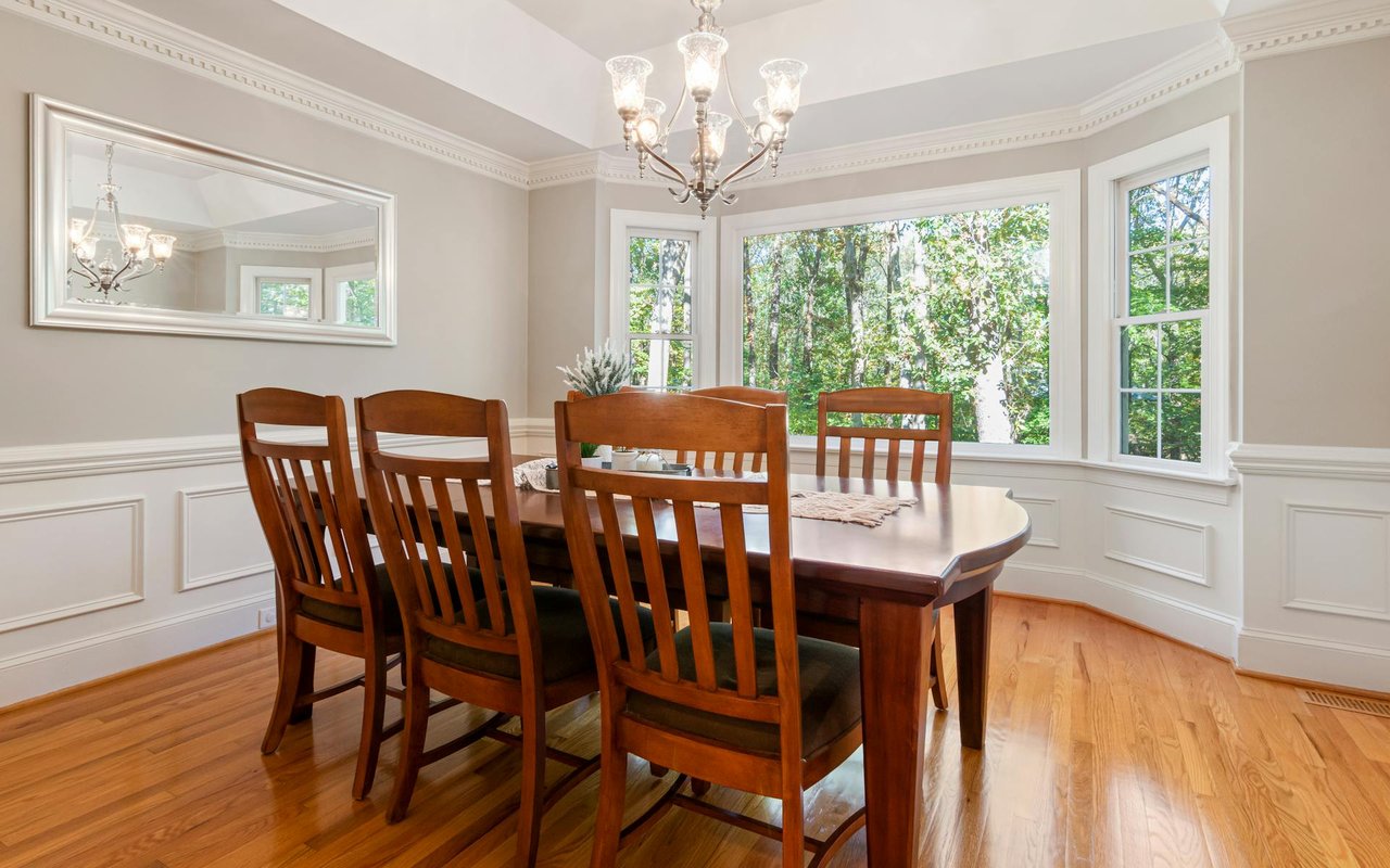 A dining room with a wooden table and chairs, a hanging chandelier, a wall-mounted mirror, and an outdoor-view window.