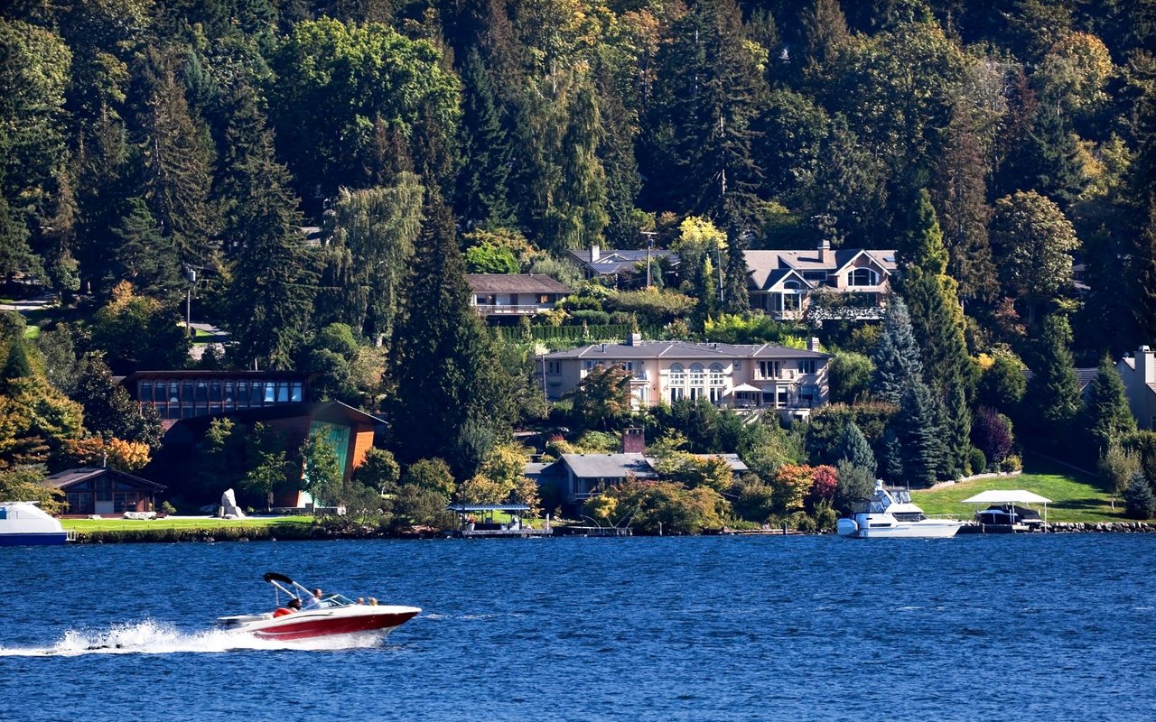 A lush green hillside is covered with dense trees and several large, luxurious houses with docks and boats along the water's edge.