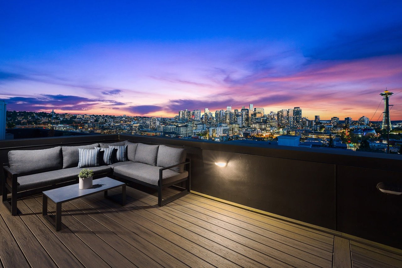 A rooftop deck with a couch and a table overlooking a city skyline at sunset.