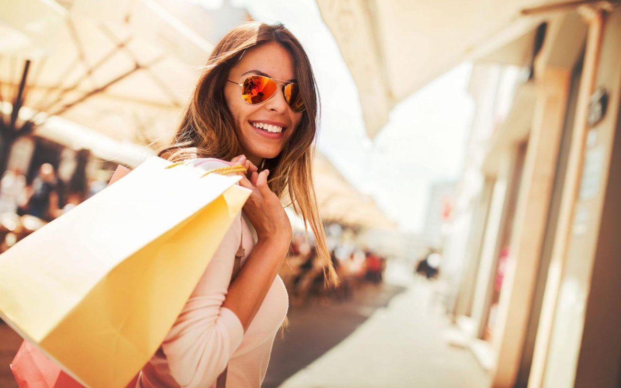  A smiling woman with long brown hair and mirrored aviator sunglasses looks over her right shoulder, carrying a large yellow shopping bag over the same shoulder.