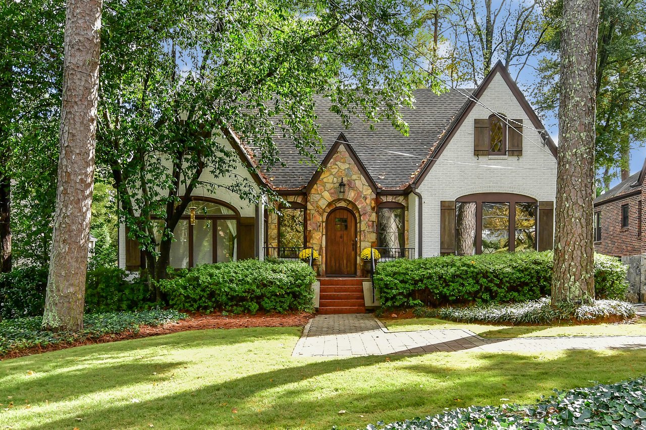 A Cape Cod cottage-style white house with a wooden door, stone roof, green lawn, and small white picket fence.